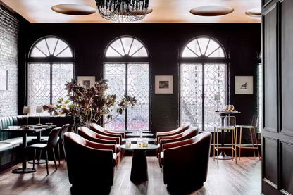 A stylish interior lounge or bar area with dark wood floors, three tall arched windows with decorative grates, brown leather armchairs, black tables, and a chandelier overhead.